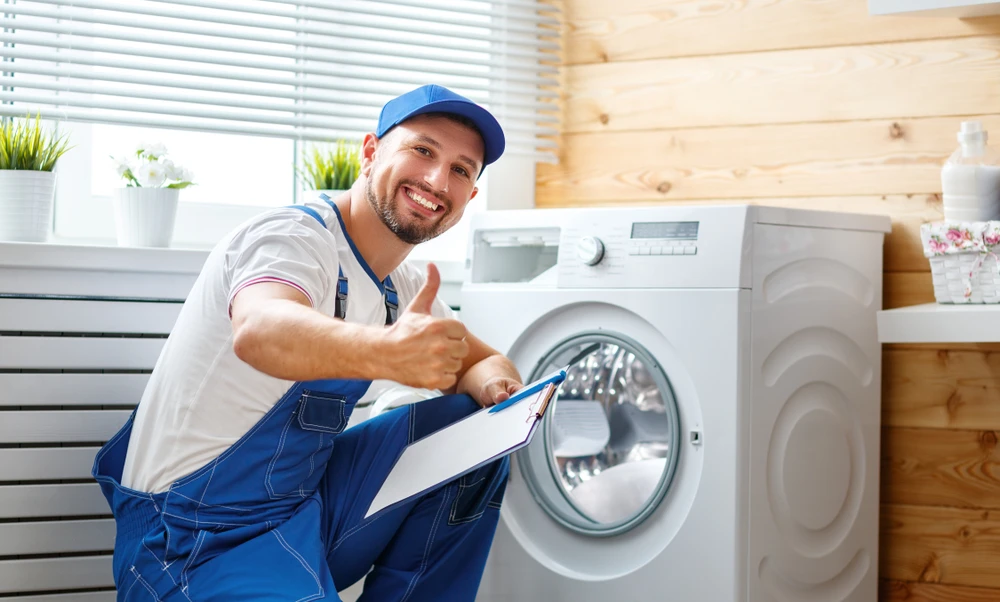 Smiling technician giving a thumbs-up while inspecting a laundry appliance, promoting the importance of Clean Your Dryer Vent Frequently.